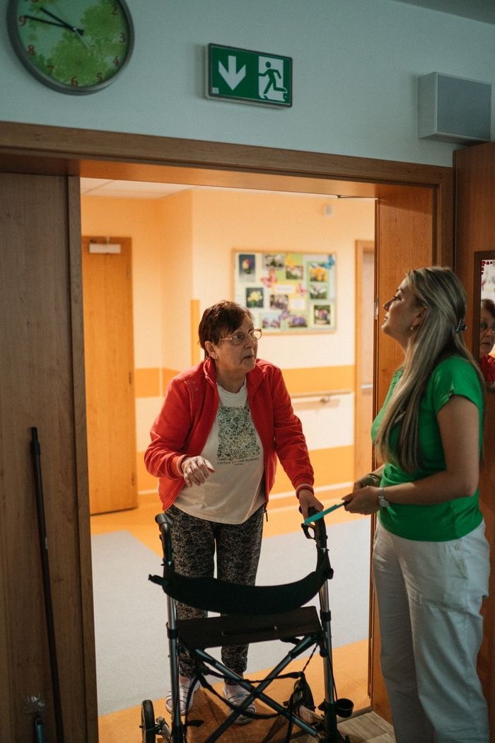 services-04 A caregiver assists a senior woman with a walker in a retirement home corridor.